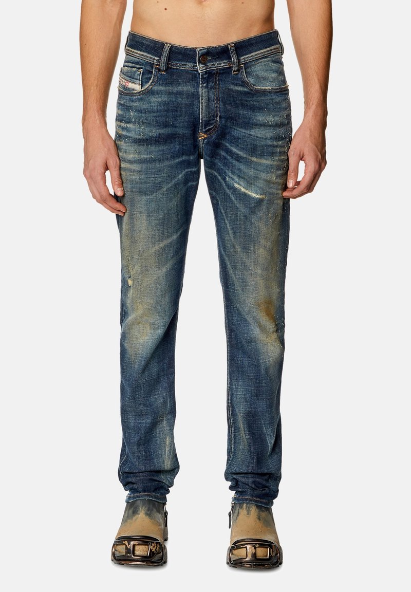Man in versleten blauwe jeans en tan laarzen met metalen teenbeschermers, staand tegen een effen achtergrond.