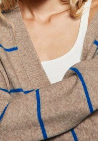 Beżowy sweter na drutach w niebieskie paski, z dekoltem w serek i miękką fakturą, częściowo zakrywający biały top na ramiączkach pod spodem.