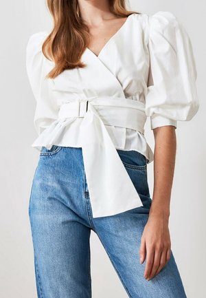 Frau trägt eine weiße Wickelbluse mit puffigen Ärmeln, einem breiten Gürtel und hoch taillierte blaue Jeans aus Denim.