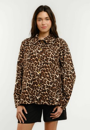 Nő hosszú ujjú, leopárdmintás gombos ingben és fekete rövidnadrágban, egy egyszerű, világos háttér előtt állva.