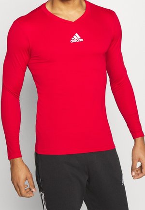 Homme portant un t-shirt rouge à manches longues Adidas avec un col en V et un pantalon noir Adidas, debout devant un fond blanc.