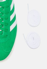 Green suede sneaker with white stripes, featuring textured laces. Includes two coiled white shoelaces laid next to the shoe.