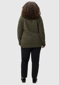 Pull vert olive avec une finition texturée, présentant de petits motifs à pois blancs. Associé à un jean noir et des baskets noires.