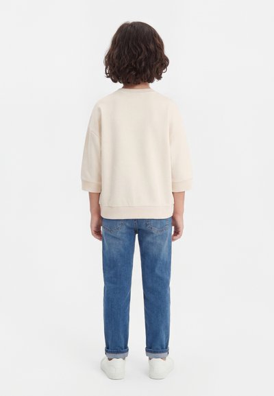 Barn med krøllet hår står med ryggen til iført en beige langærmet trøje, blå jeans med manchetter og hvide sneakers på en hvid baggrund.