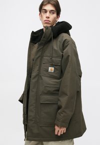 Parka di colore verde oliva con cappuccio foderato in pile nero, tasche frontali e patch con logo. Presenta una chiusura con zip e bottoni a pressione.