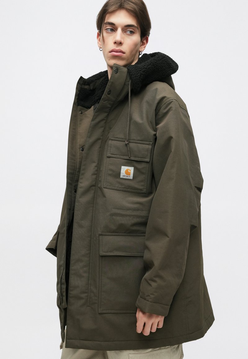 Parka di colore verde oliva con cappuccio foderato in pile nero, tasche frontali e patch con logo. Presenta una chiusura con zip e bottoni a pressione.