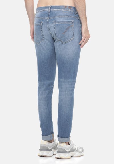 Uomo che indossa jeans azzurri arrotolati e sneaker bianche, in piedi con la schiena rivolta verso la camera su uno sfondo neutro.