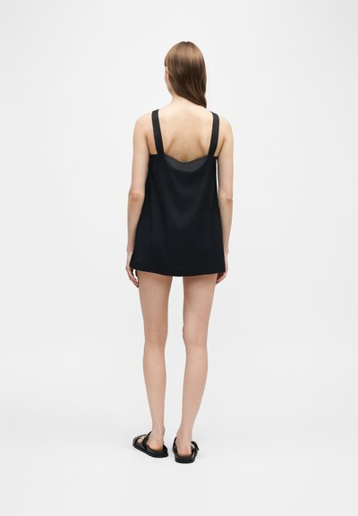 Femme debout, de dos, portant une robe noire courte sans manches avec de larges bretelles et des sandales noires sur un fond clair uni.