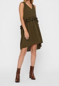 Oliivinvihreä hihaton mekko, jossa on röyhelöt yksityiskohtina vyötäröllä, V-pääntie ja epäsymmetrinen helma. Ruskeat nilkkurit.