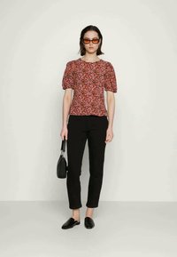 Top floral de mangas bufantes em rosa e roxo numa modelo, combinado com calças pretas ajustadas e sapatos pretos tipo mocassim, segurando uma pequena bolsa preta.