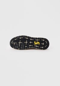 Schwarze Gummisohle mit strukturiertem Muster und beigen Akzenten, ausgestattet mit einem gelben Logo und detaillierten Rillen für besseren Grip.