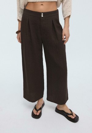 Person trägt hoch taillierte, weit geschnittene dunkelbraune Hose, beigen Cropped-Pullover, schwarze Plateau-Zehensandalen und ein braunes Armband am linken Handgelenk.