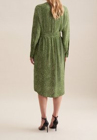 Zelené šaty s jemným vzorem bílých teček, dlouhými rukávy a úzkým pasem. Límec končí u poloviny lýtek. Nosené s černými sandály na vysokém podpatku.