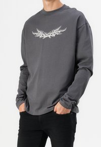 Homme portant une chemise à manches longues gris foncé avec un motif tribal abstrait blanc sur la poitrine et un pantalon noir, mains dans les poches.