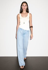 Top canotta giallo chiaro, jeans larghi blu chiaro, braccialetto marrone sul braccio sinistro. Il modello cammina su un pavimento scuro contro uno sfondo bianco semplice.