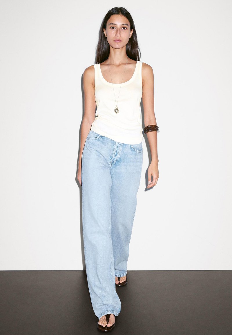 Top canotta giallo chiaro, jeans larghi blu chiaro, braccialetto marrone sul braccio sinistro. Il modello cammina su un pavimento scuro contro uno sfondo bianco semplice.