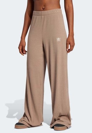 Osoba nosząca beżowe, szerokie, prążkowane spodnie z logo Adidas w pobliżu talii, w połączeniu z czarnymi i brązowymi butami, stojąca na jednolitym tle.