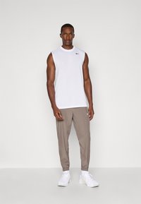 Hvid ærmeløs sportsbluse med et sort logo, parret med lysebrune tapered joggers og hvide sportsko.