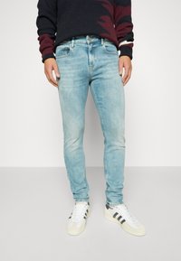 Vybledlé světle modré skinny džíny s hladkou texturou, doplněné bílými sportovními teniskami s černými pruhy.