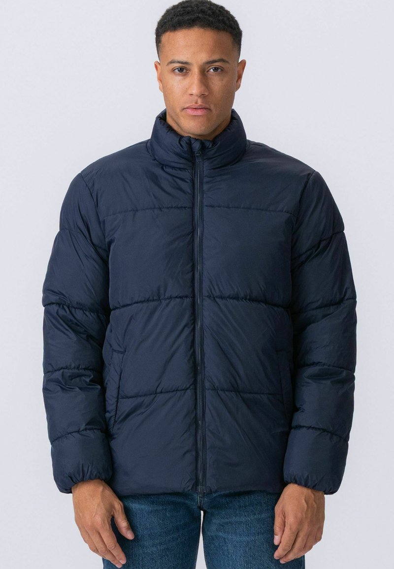 Moški, ki nosi temno modro podloženo jakno z zadrgo, stoji ob preprostem svetlem ozadju z rokami sproščeno ob telesu.