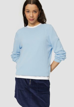 Pull en maille bleu clair avec col rond et manches longues, superposé sur une chemise blanche, associé à une jupe en corduroy bleu marine.