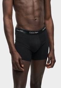 Αρσενικός κορμός και άνω πόδια που φορούν μαύρα boxer Calvin Klein με λογότυπο στη μέση σε απλό ανοιχτό φόντο.