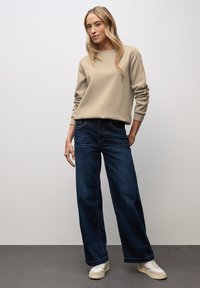 Beżowa bluza z okrągłym dekoltem, ciemnoniebieskie szerokie dżinsy i białe sneakersy. Gładka faktura, luźny krój, z widocznymi detalami szwów.