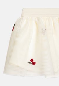 Jupe en tulle crème avec une taille élastique. Présente une broderie de cerises rouges et une texture douce et transparente pour un aspect superposé.