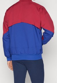 Färgblockerad jacka med en röd övre del och en blå nedre del. Tillverkad av mjukt tyg, med en ribbad midja och muddar.