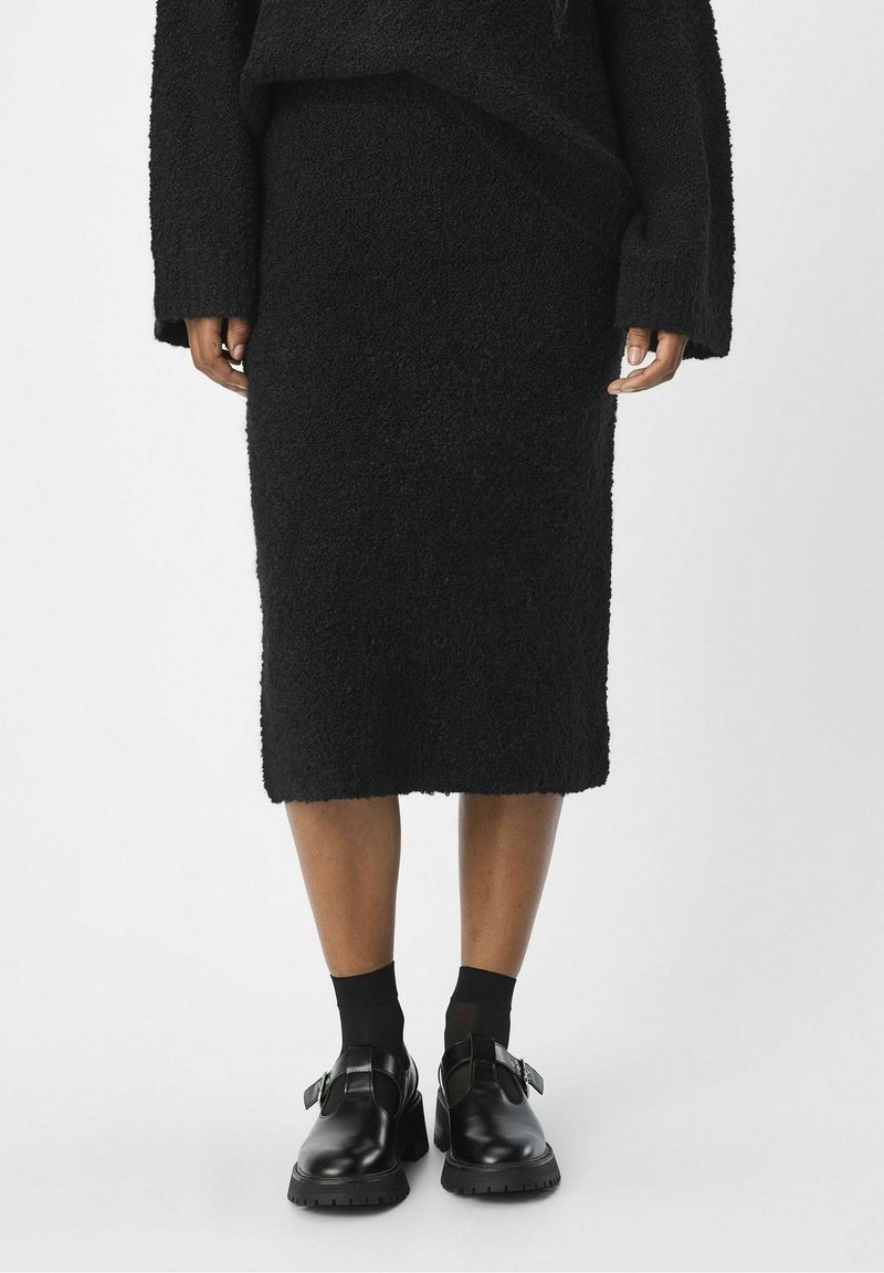 Pull noir en tricot et jupe crayon noire assortie avec une surface texturée, accompagnés de chaussures noires avec des accents à boucle et des semelles épaisses.