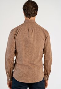 Bruin shirt met lange mouwen van een gestructureerde stof. Voorzien van knoopmanchetten en een plooi aan de achterkant voor meer bewegingsvrijheid. Gladde, subtiele afwerking.