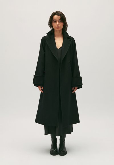 Schwarzer Wollmantel mit lockerer Passform, großem Kragen und breiten Manschetten mit Knopfdetails. Trägt einen schwarzen Kleid und kombiniert mit schwarzen Stiefeln.