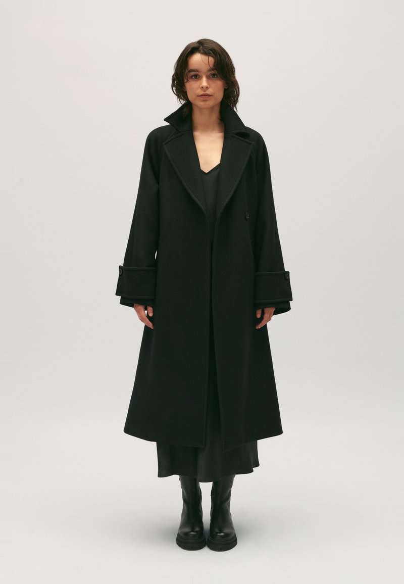 Zwarte wollen coat met een relaxed fit, grote kraag en brede mouwen met knopdetails. G gedragen over een zwarte jurk en gecombineerd met zwarte laarzen.