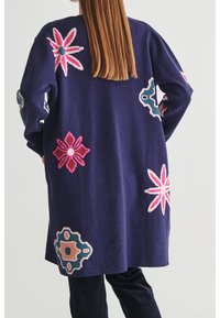 Dlouhý námořnický kardigan s živými květinovými vzory v růžové, červené a tyrkysové barvě, vyrobený z měkké tkaniny, s volným, uvolněným střihem.