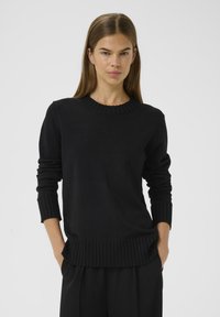 Eine Frau mit glattem braunen Haar, die einen schwarzen gerippten Pullover und schwarze Hose trägt, steht mit den Händen in den Taschen vor einem schlichten Hintergrund.