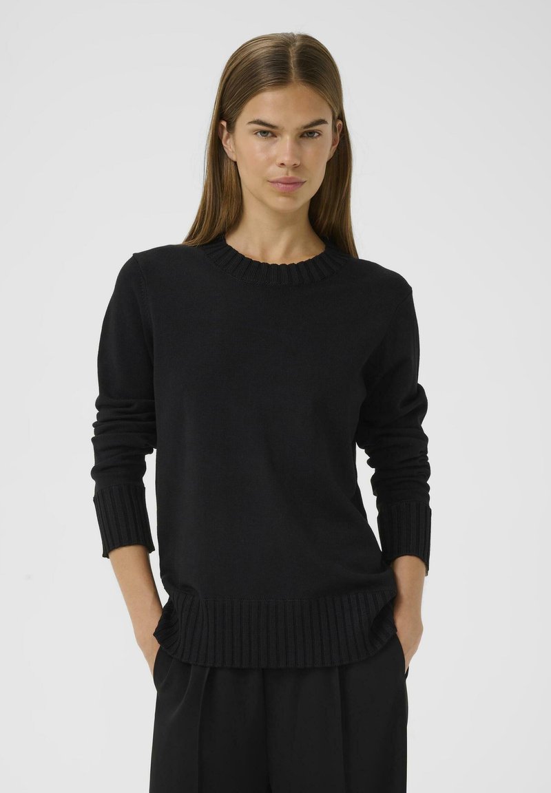 Eine Frau mit glattem braunen Haar, die einen schwarzen gerippten Pullover und schwarze Hose trägt, steht mit den Händen in den Taschen vor einem schlichten Hintergrund.