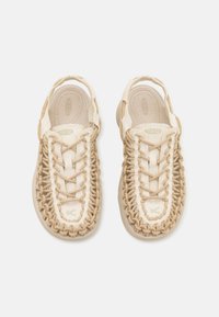 Chaussures beige avec une tige en textile tissé et une semelle en caoutchouc. Elles présentent des lacets et un design texturé avec un logo en accent vert.