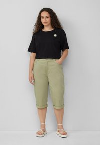 Zwarte katoenen T-shirt met zon-graphics, gecombineerd met lichtgroene knielange broek en strap sandalen in het wit, staande tegen een effen achtergrond.