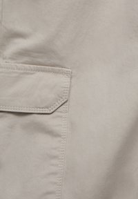 Gros plan sur un tissu beige avec une poche rectangulaire cousue comportant un rabat aux coins biseautés en haut à gauche.
