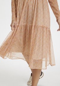 Lång, genomskinlig, blommig klänning i ljus persika med rosa och gula detaljer. Har ett lager på lager nederdel och långa ärmar, kombinerad med sneakers.