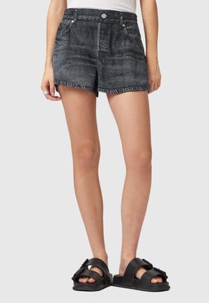 Kvinde iført sorte vasket denimshorts og sorte sandaler med dobbelte stropper, stående foran en ensartet lys baggrund.