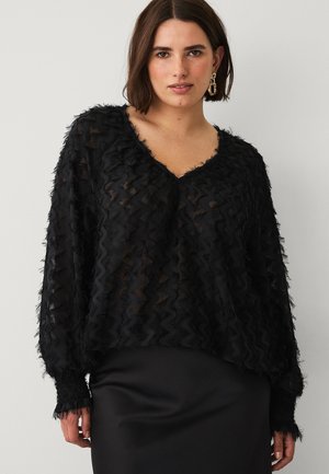 Vrouw met kortbruin haar, gekleed in een zwart doorschijnend blouse met een golvend patroon en gouden creolen, tegen een effen achtergrond.