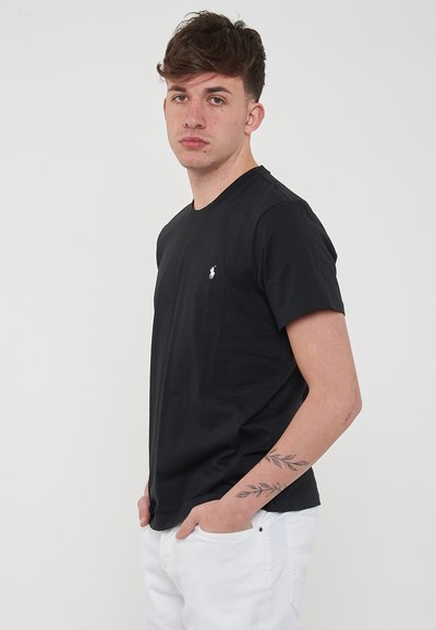 T-shirt nera in cotone con scollo tondo e maniche corte, con un piccolo logo bianco sul lato sinistro. Indossata con pantaloni bianchi.