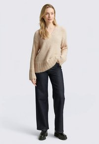 Lichtbeige trui met V-hals gemaakt van zachte, gestructureerde stof met geribbelde manchetten, gecombineerd met donkerblauwe wijde jeans en zwarte laarzen.