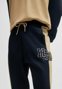 Joggers navy e beige realizzati in tessuto morbido, dotati di un cordoncino regolabile, logo ricamato e un sorriso stilizzato sul lato.