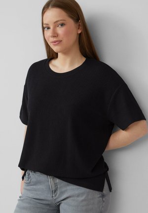 Zwarte gebreide t-shirt met korte mouwen, ronde halslijn en zijsplitten. De textuur is geribbeld en het is gecombineerd met lichtgrijze jeans.
