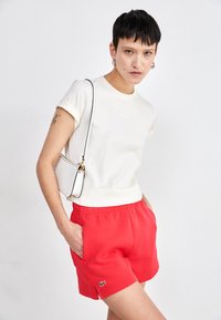 T-shirt a maniche corte color crema con logo in rilievo, abbinato a pantaloni corti rossi con vita elasticizzata, caratterizzati da tasche laterali e un emblema Lacoste.