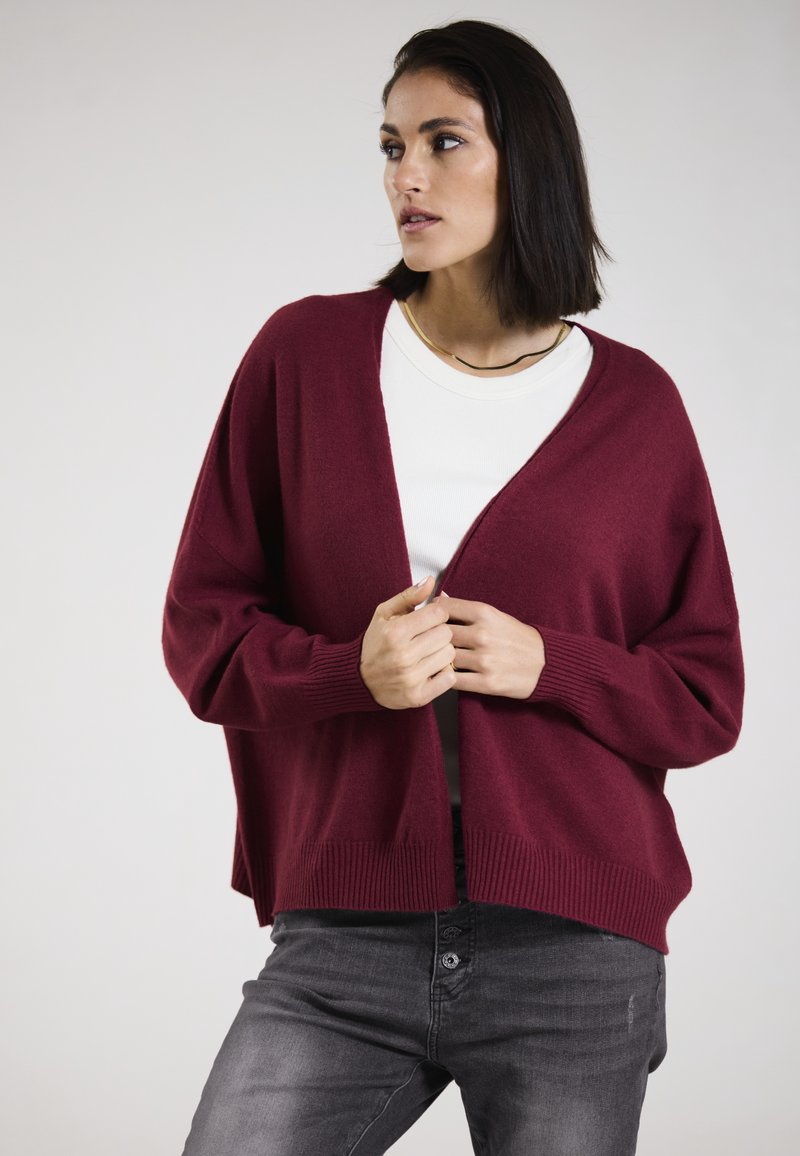 Bordeauxfarbener Cardigan aus weichem Material mit lockerer Passform, offenem Vorderteil und gerippten Bündchen; kombiniert mit einem weißen Oberteil und dunklen grauen Jeans.