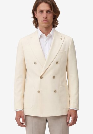 Mann mit mittellangen braunen Haaren, der einen cremefarbenen Doppelreiher-Blazer über einem weißen Hemd und hellen Hosen trägt.
