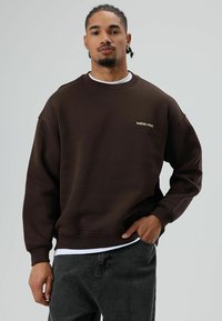 Bruine trui met een ronde hals en verlaagde schouders, met een klein geel geborduurd tekstontwerp op de borst. Gecombineerd met donkere jeans.